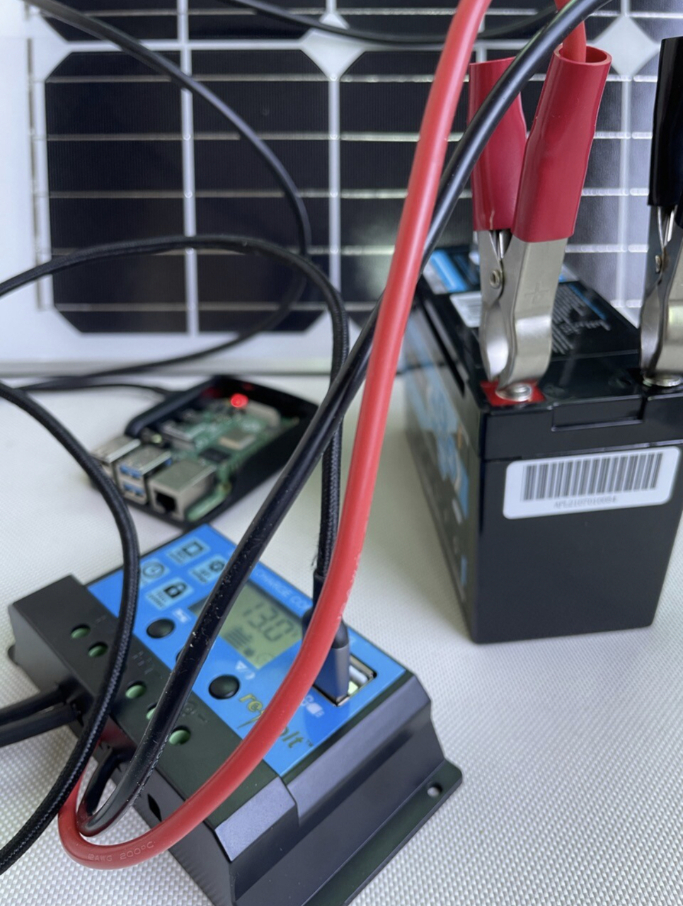 An electronic setup with a solar panel, a car battery, a little device called MPPT that regulates the power coming from the solar panel and a raspberry pi. All four are connected with red and black cables. The display of the MPPT shows 13V and the raspberry pi has a red LED lit up.